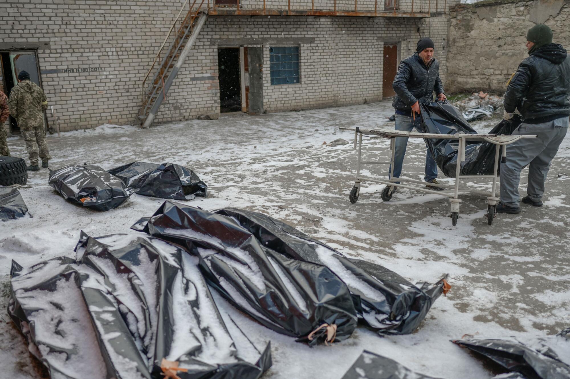 Two men carry a corpse in a body bag to lay it next to others in a snow covered yard in Mykolaiv.
