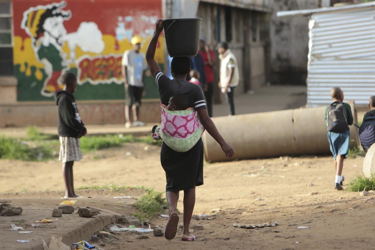 A woman walks carrying her baby in a back sling and holding a bucket of water steady on her head