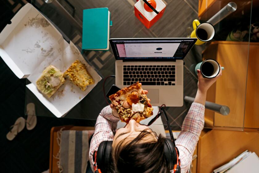 A middle-aged woman sitting in the kitchen at the glass table with laptop in headphones and holding pizza and coffee.