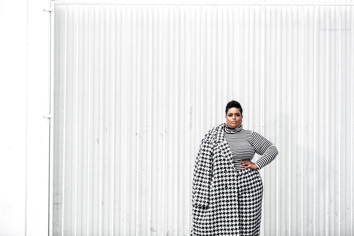 A woman in houndstooth check pants and coat stands in front of a white backdrop.