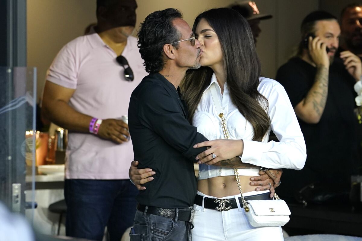 A middle-aged man holds and kisses a younger woman in a luxury box before a sporting event.