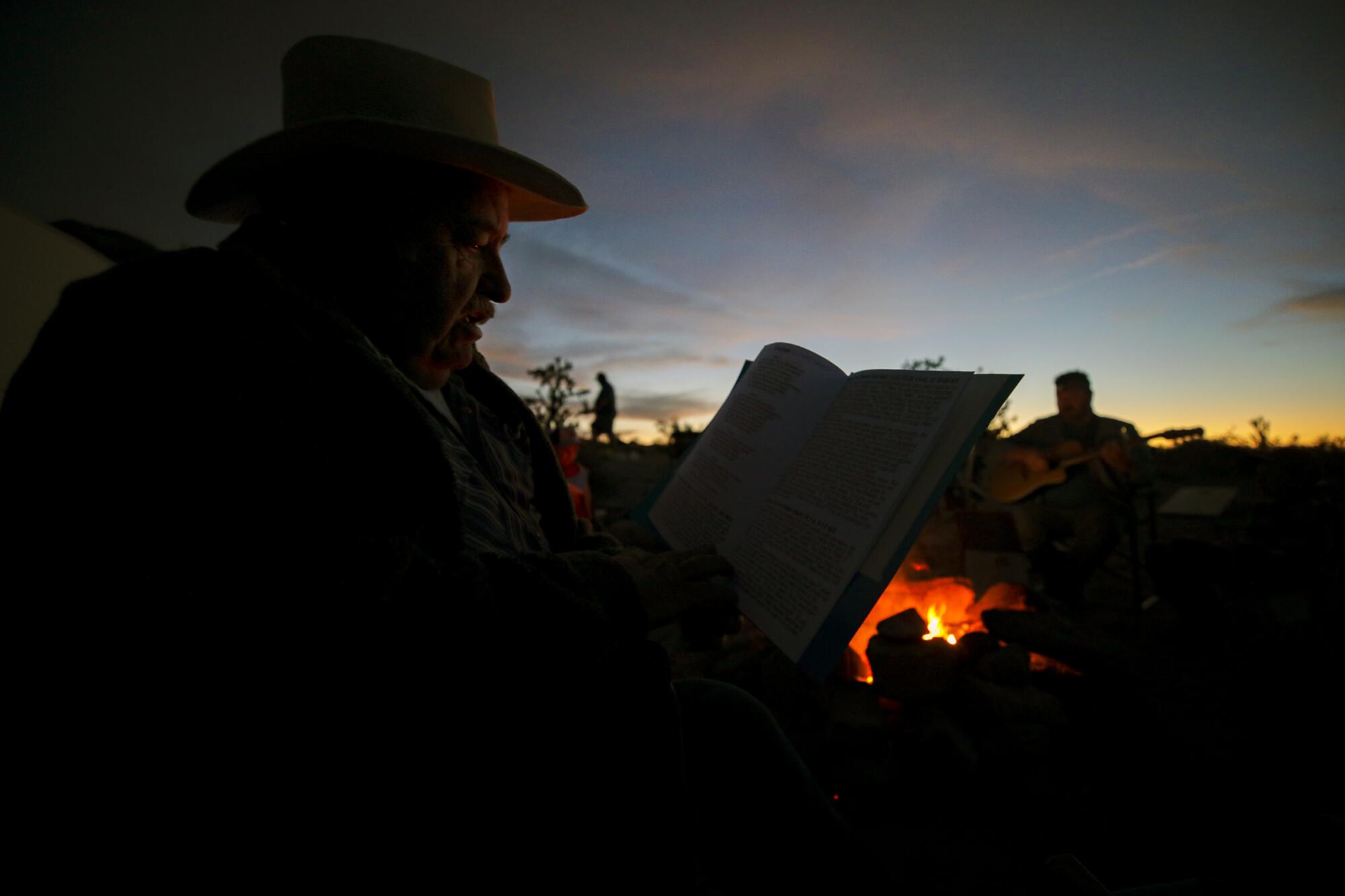 A man reads pages, silhouetted against the dawn sky with a campfire in the background
