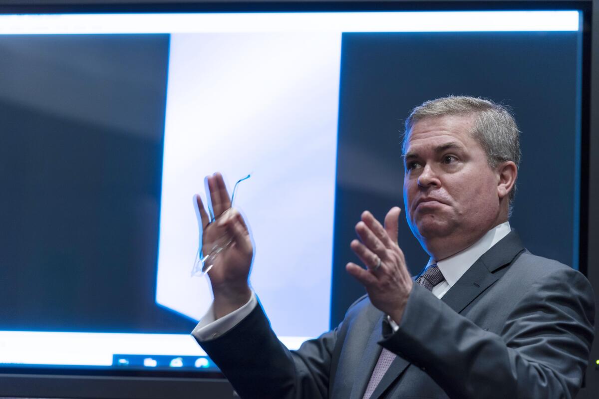 A man in a gray suit holds up his hands in front of a video display of a UAP.
