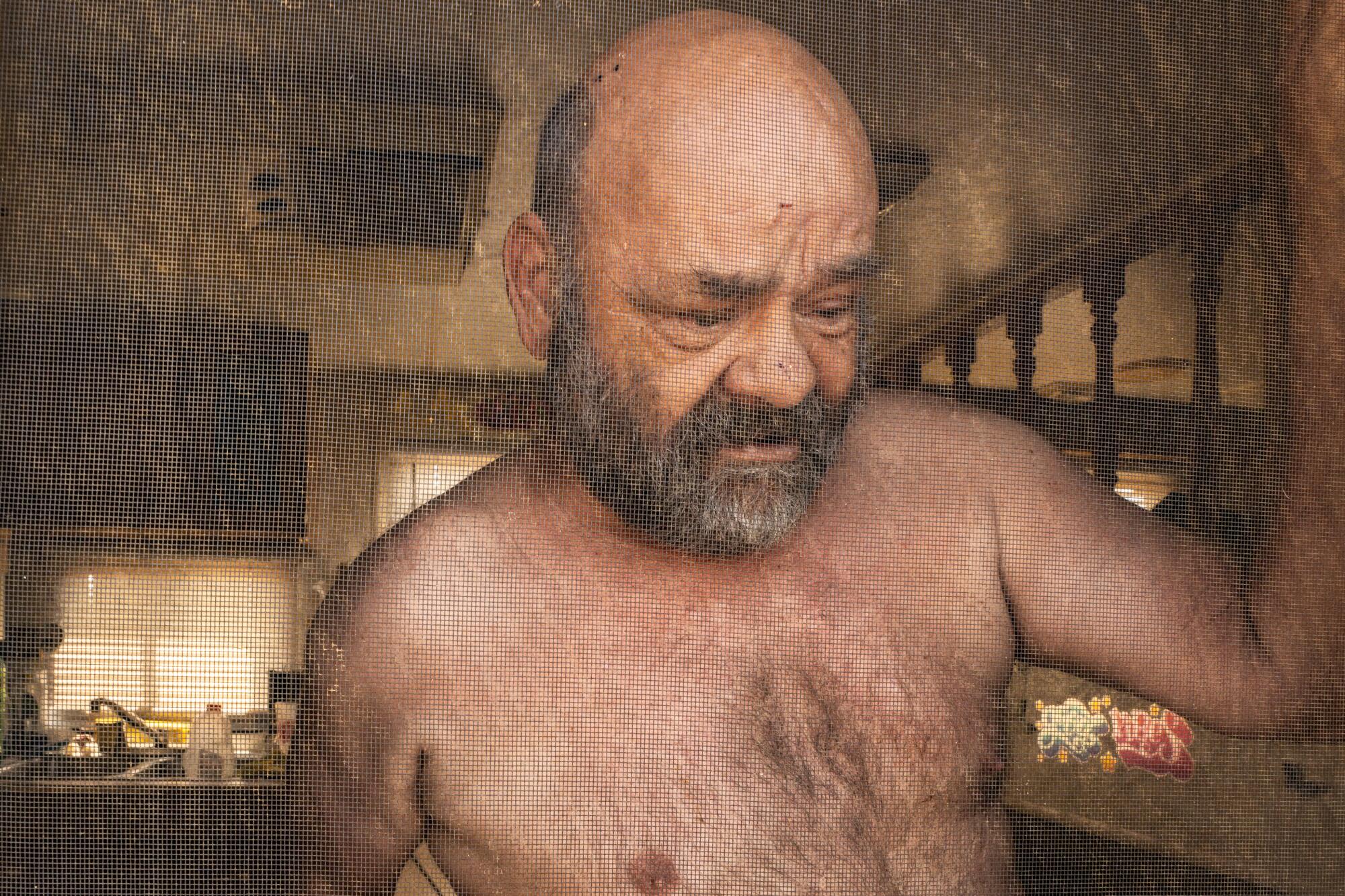 A shirtless man with a beard holds up one arm. 