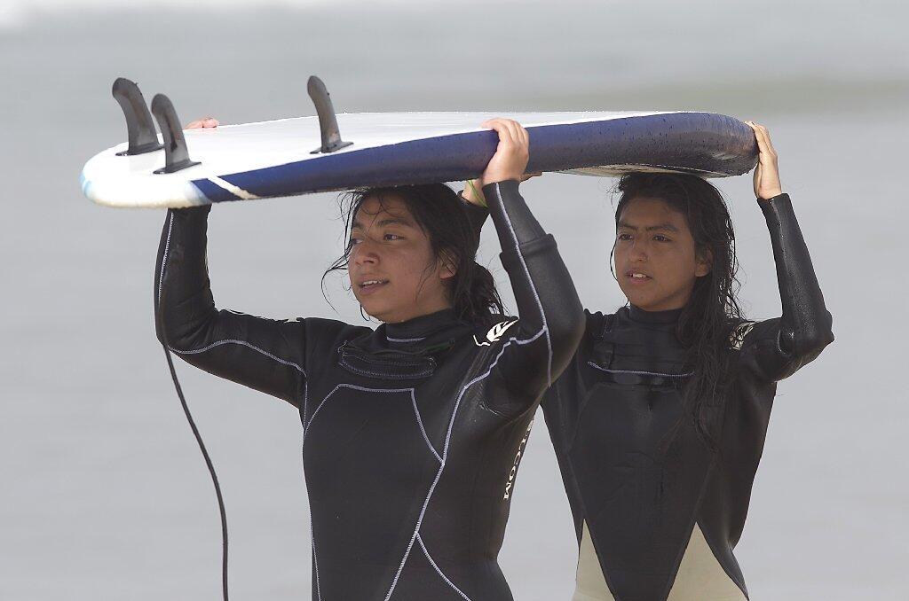 Surfers Jackie Olivares, left, and Anette Mongers carry a board together during Save Our Youth’s Surf Days program in west Newport on Thursday.