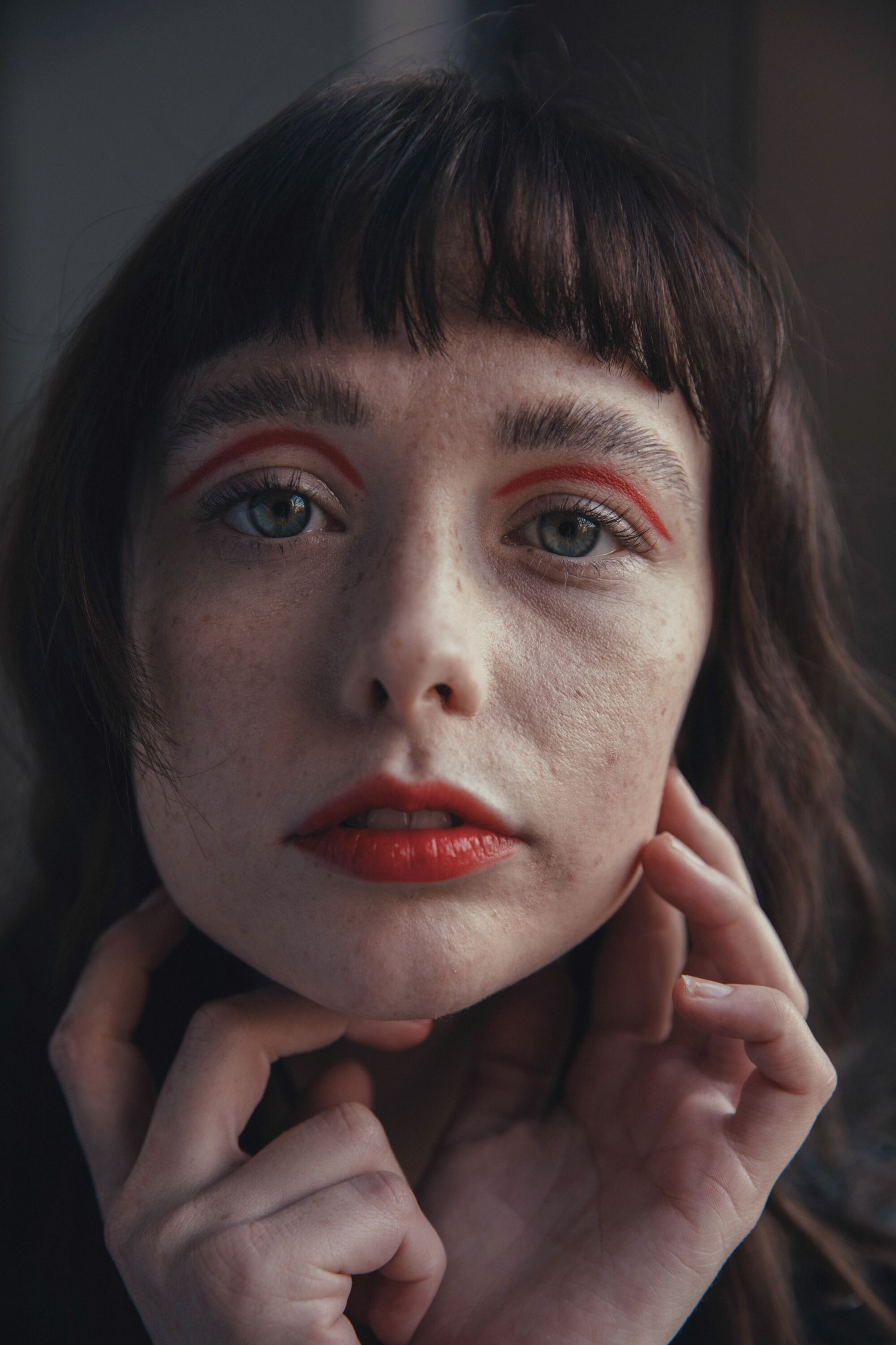A woman with red lipstick and red eye makeup holds her hands to her face.