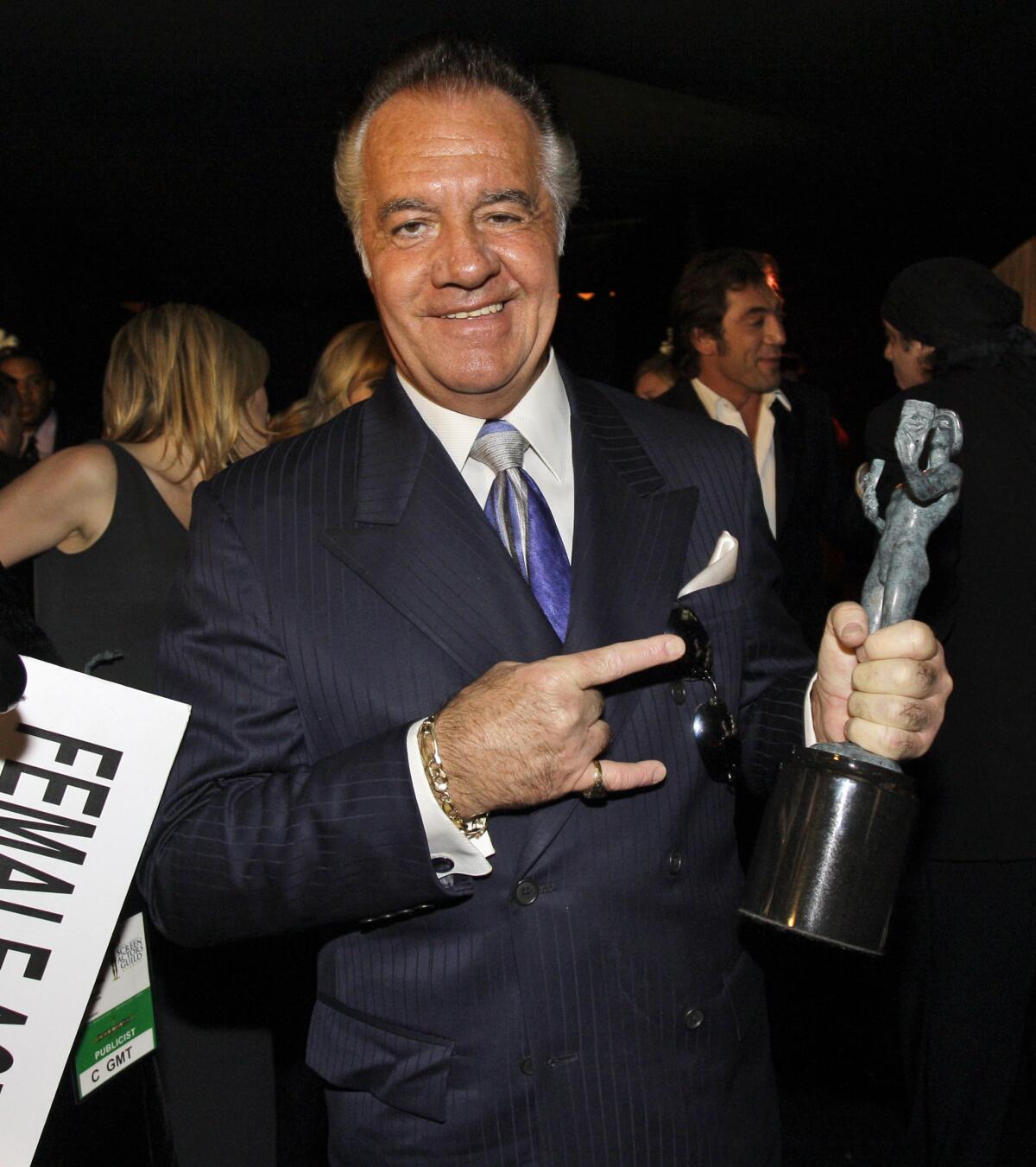 A man in a suit gestures with one hand toward the award statue he holds in the other hand