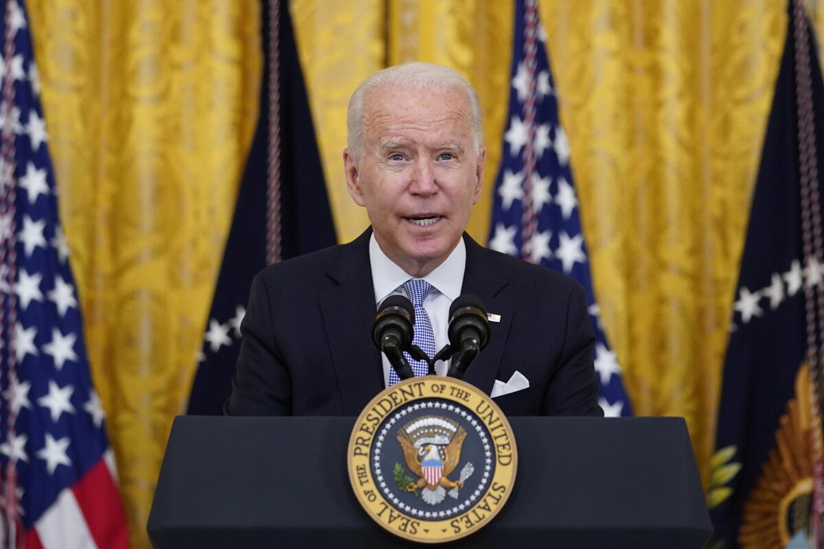 El presidente Biden habla desde un atril con el sello presidencial sobre un fondo de banderas y cortinas de color amarillo dorado.