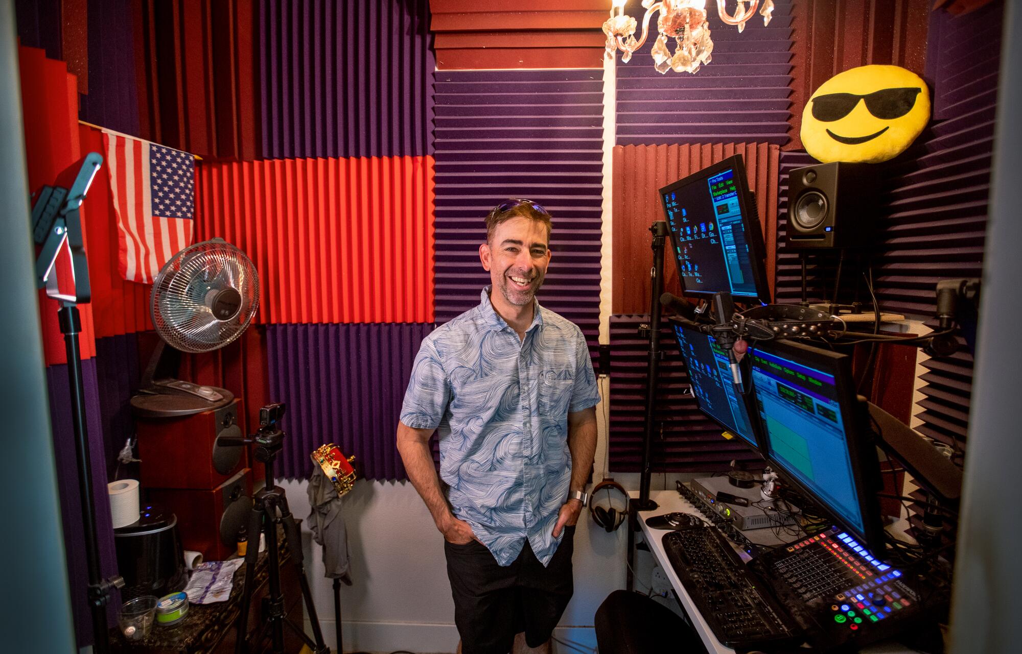 A man stands in a room with purple and red foam acoustic panels with computer screens and sound equipment around him.