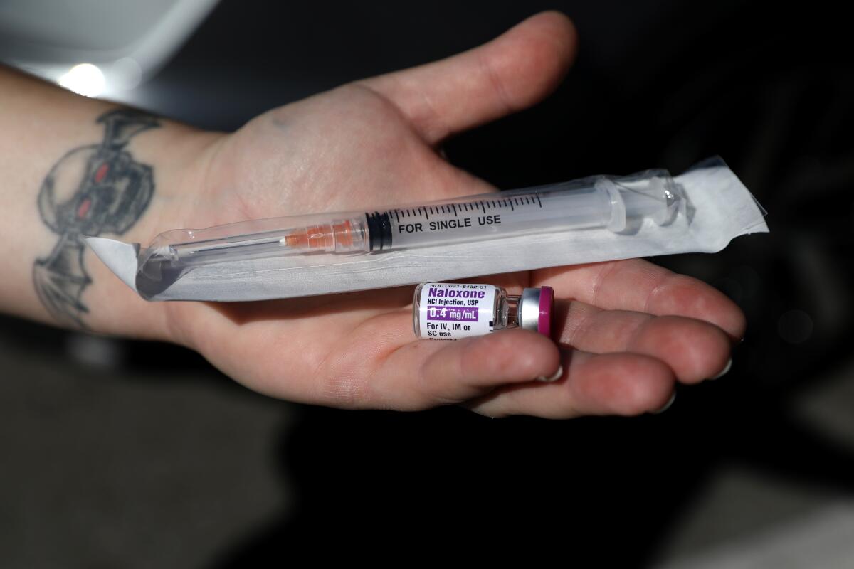 A hand is shown holding a small vial of naloxone and a syringe in a sterile package. 