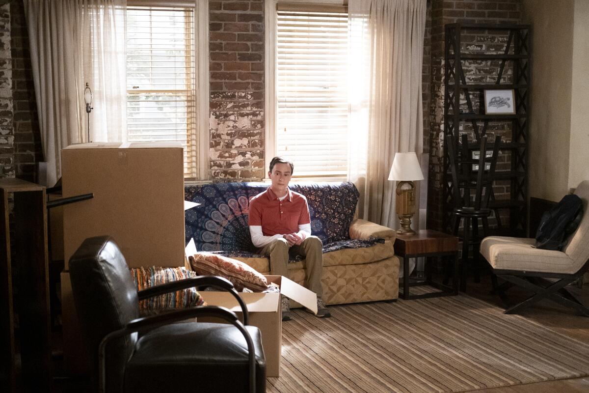 A young man sits on the couch in his apartment, surrounded by moving boxes