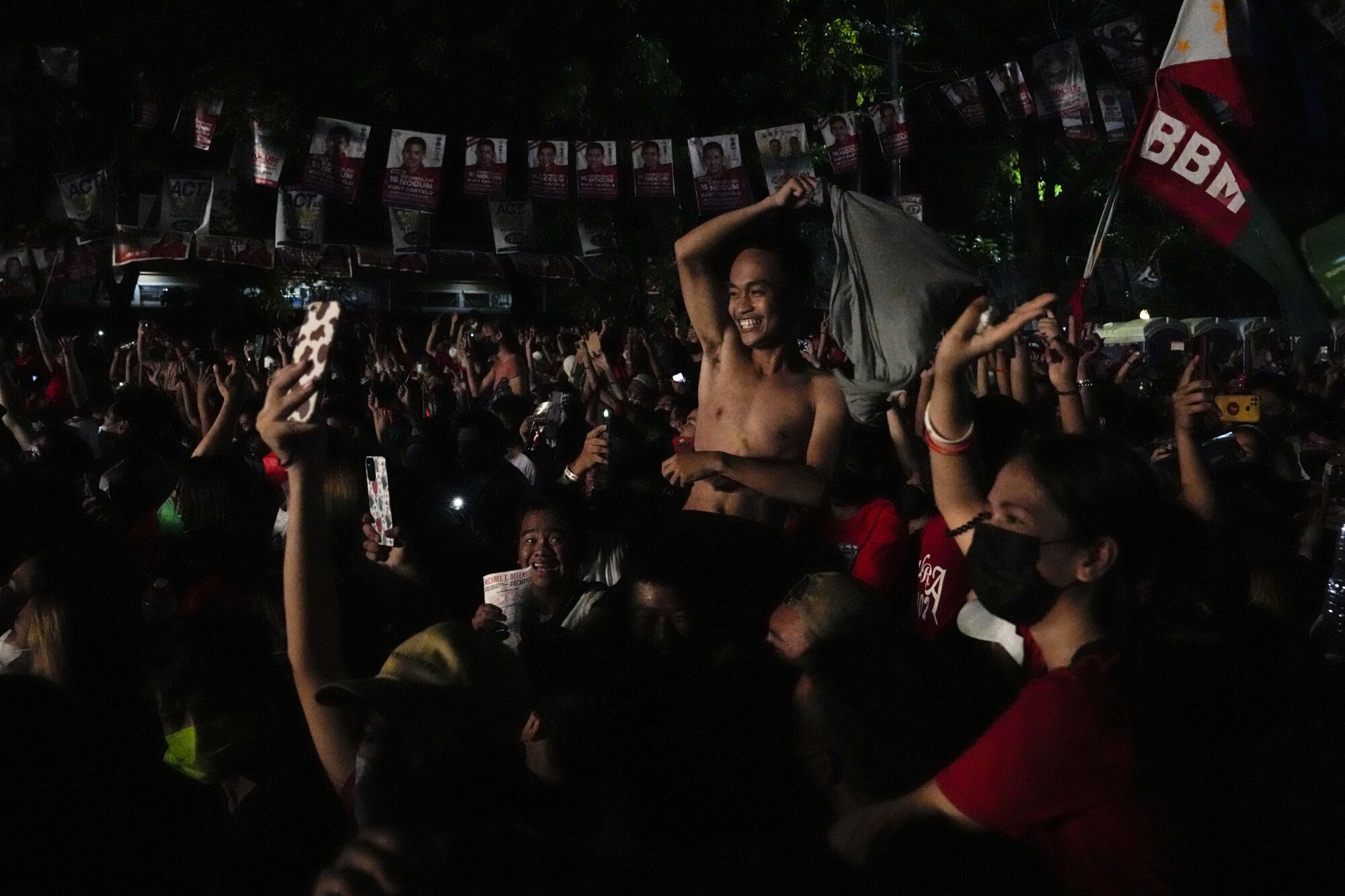 A shirtless man smiles, raising one arm, as he is surrounded by other people with campaign posters overhead behind him