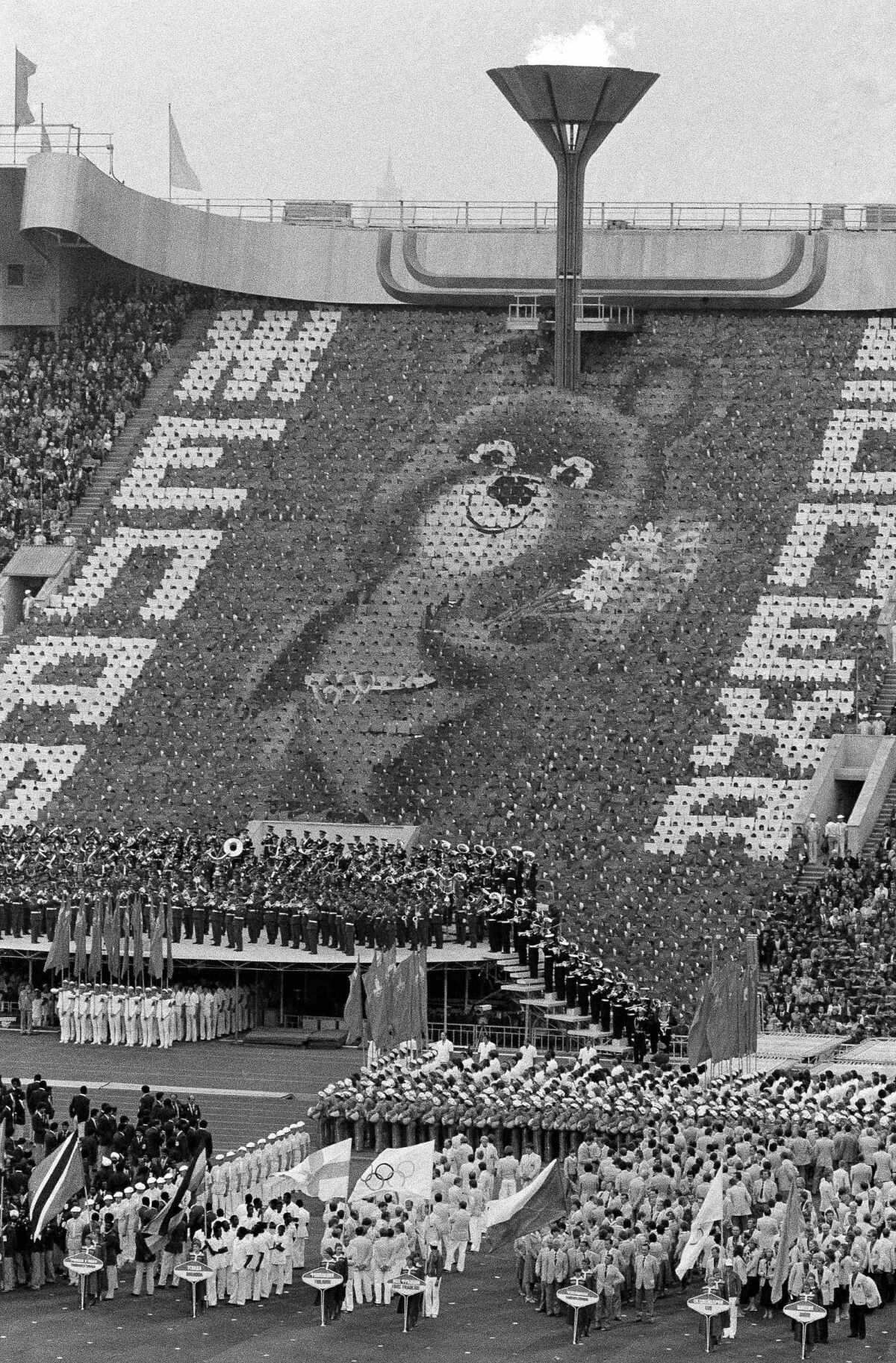 FILE - In this July 19, 1980, file photo, a giant image of Misha, the Russian bear mascot of the 1980 Moscow Summer Olympic Games, greets athletes standing on the field of Moscow's Lenin Stadium during opening ceremonies of the competitions in Moscow. The image was created by 3,500 card-bearers. Overhead burns the Olympic flame. (AP Photo/File)