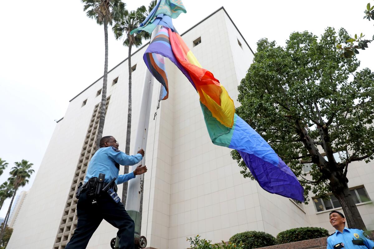 A uniformed man raises the Pride flag outside a building as another uniformed man watches.