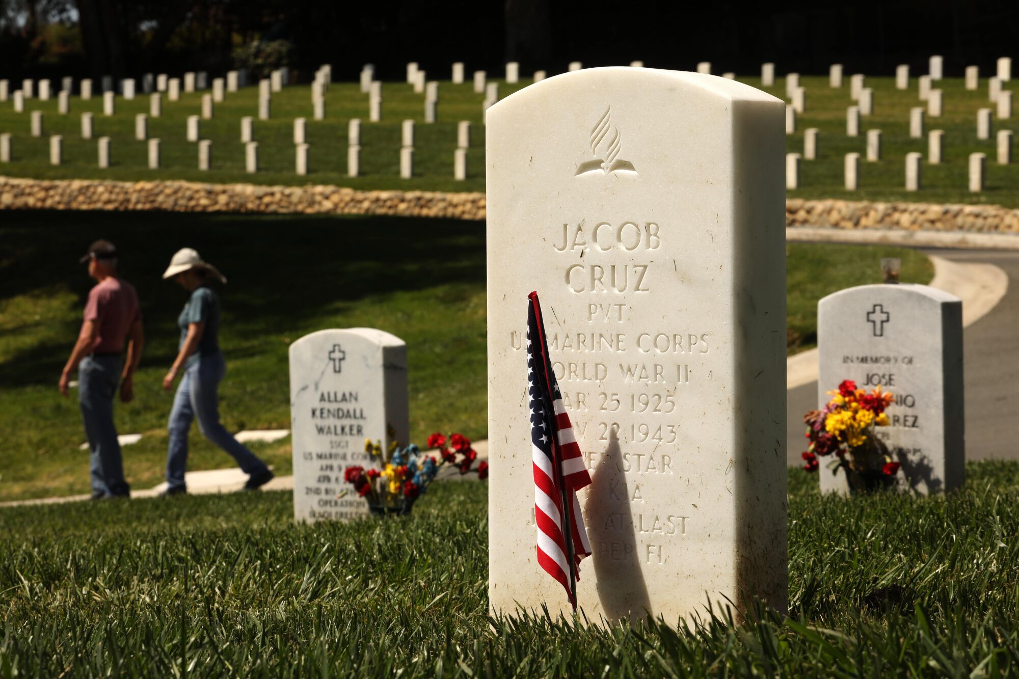 A tombstone with an American flag planted in front of it sits in a cemetery.