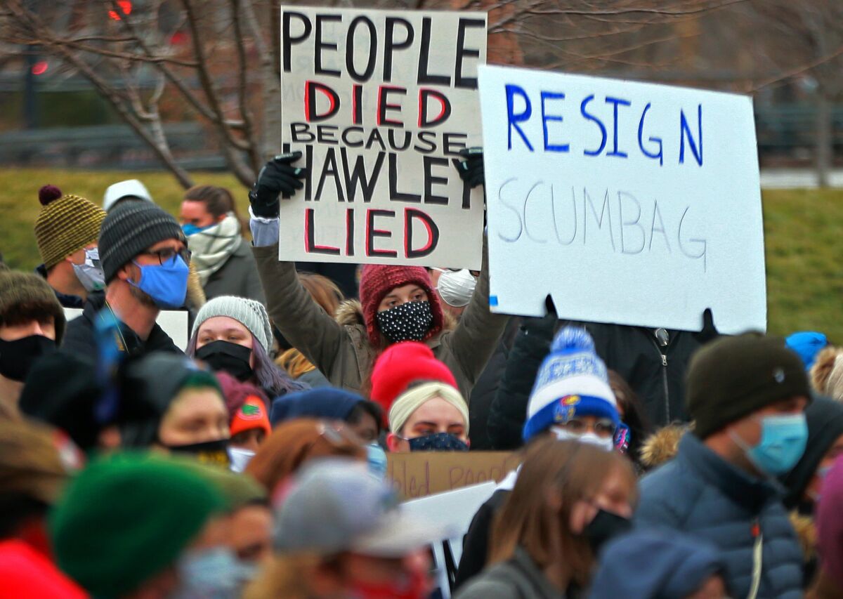 Demonstrators gather with signs saying "People Died Because Hawley Lied" and "Resign Scumbag" outside. 