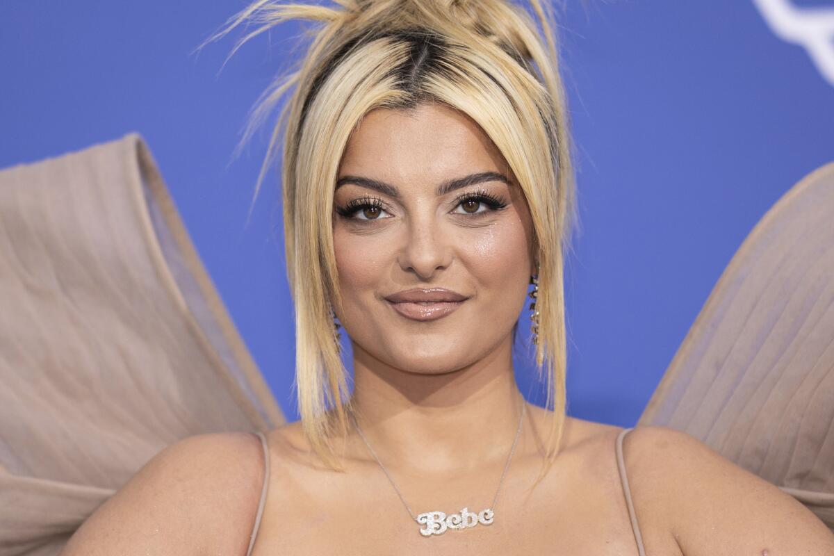 Bebe Rexha poses in a beige gown against a blue backdrop with a messy updo 