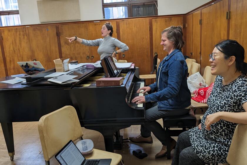 Three people around a piano watch a rehearsal.