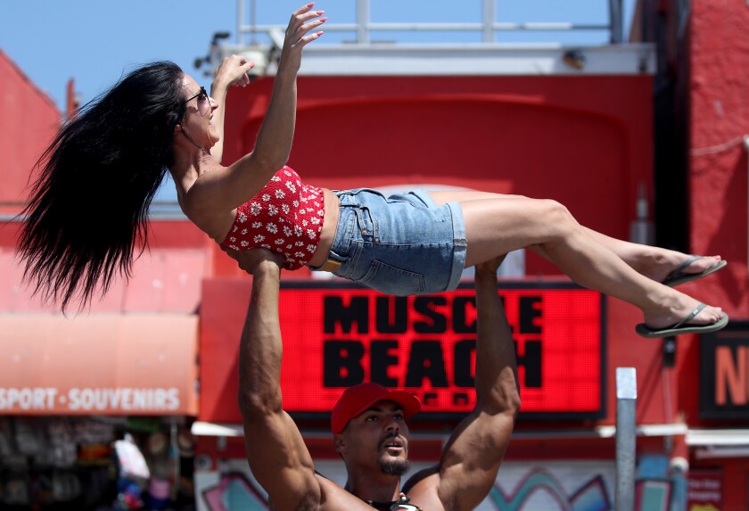 A man raises a woman above his head. In the back is a sign that says "Muscle Beach."