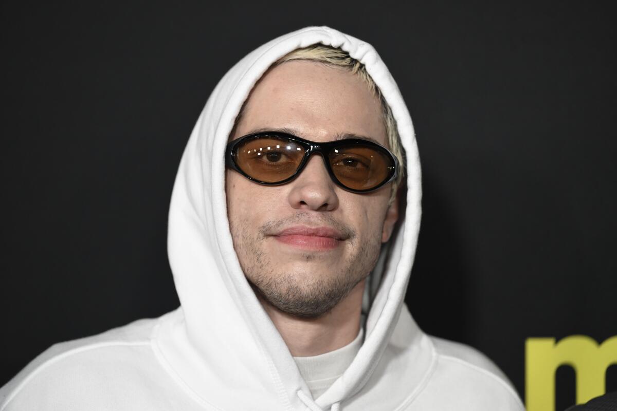 Pete Davidson wearing a white hoodie and black sunglasses with amber lenses, giving a half smile.