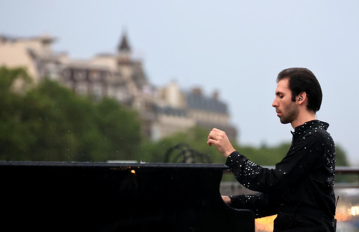 A man in black plays a piano outdoors, large chateau-like buildings visible in the distance