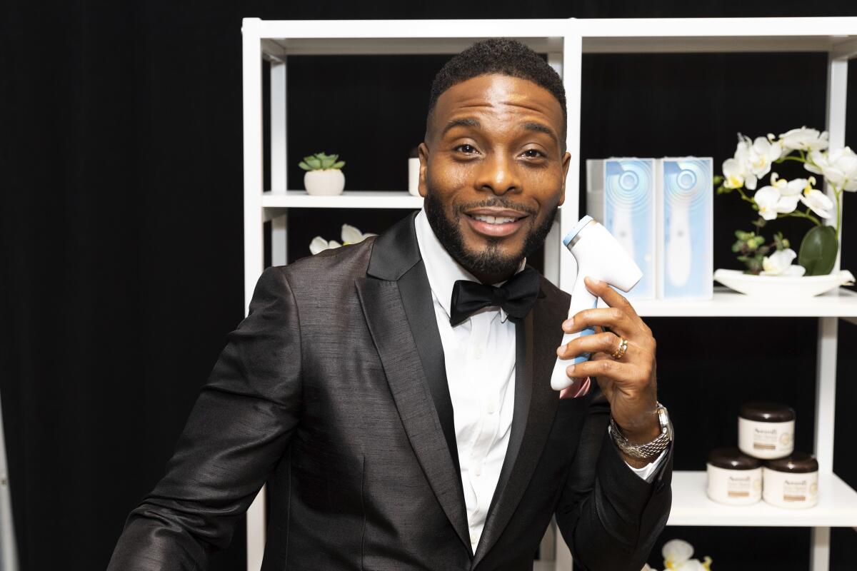 Kel Mitchell holds a white device to his face while smiling in a black and white tuxedo