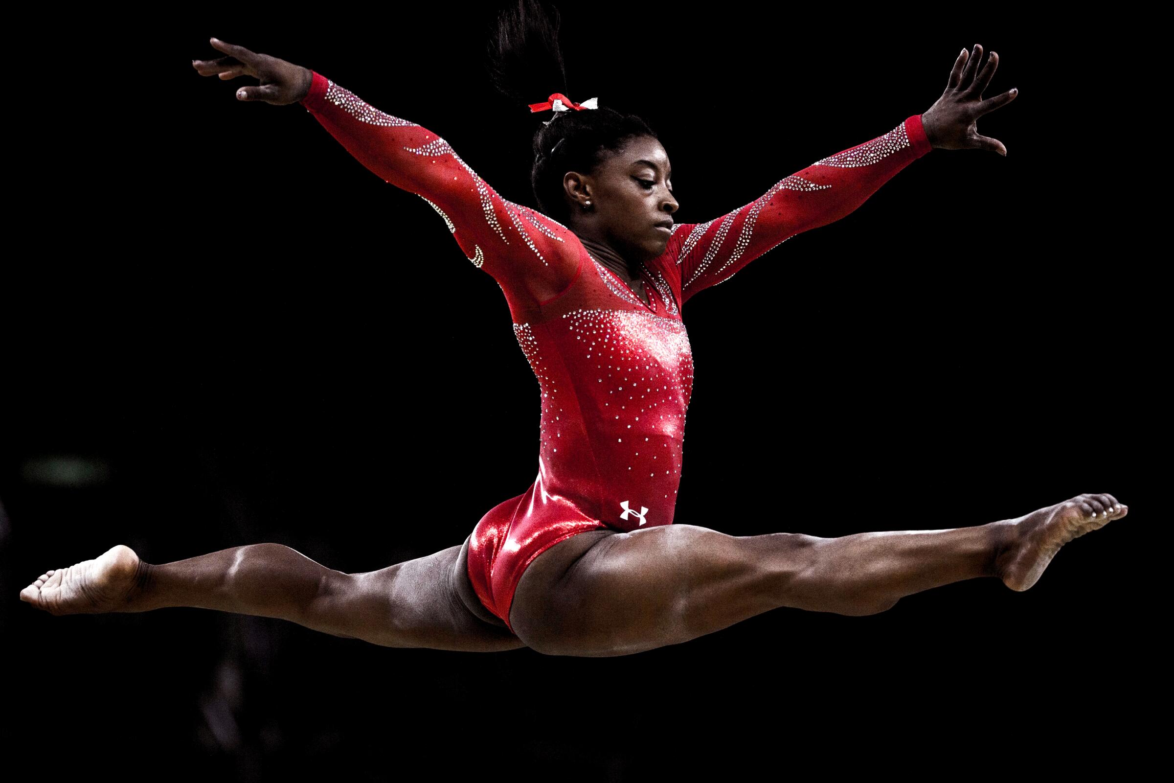 Simone Biles in a red, long-sleeve leotard leaping in the air with arms extended.