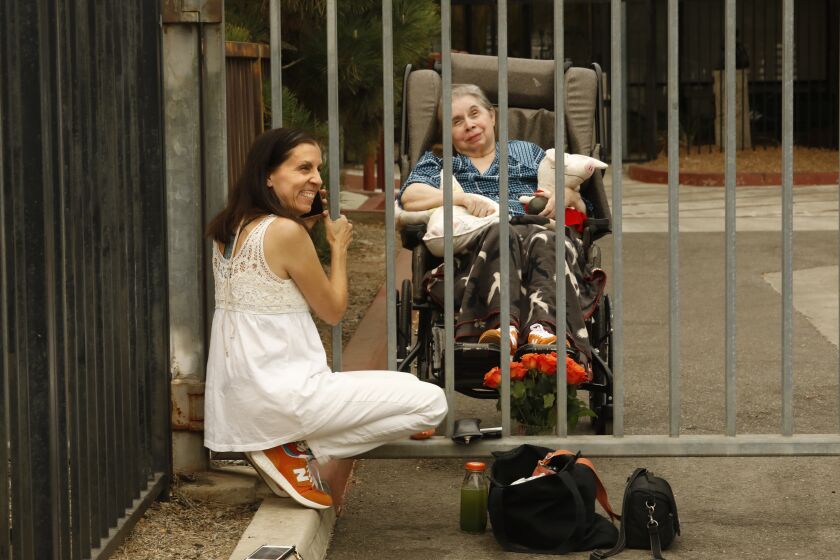 Reseda, California-Sept. 11, 2020-Photographer/writer, Hannah Kozak, age 60, visits her mother, Rachel Dietsch, 82, at the gate of the nursing home where Rachel lives. Rachel has been disabled since 1974, after her second husband hit her so hard she sustained catastrophic head injuries. He was never charged with a crime. Hannah has just published a book of photos about her mother and their family trauma, "He Threw the Last Punch Too Hard." Rachel blamed her mother for the disintegration of their family-for leaving her father and five kids to marry a charming abuser. She did not see her mother for 35 years, as her mom lived in a terrible nursing home. She finally reconnected and sought to heal by photographing her mother, and coming to appreciate her sweetness and love. Hannah visits Rachel at the gate because of COVID-19. (Carolyn Cole/Los Angeles Times)