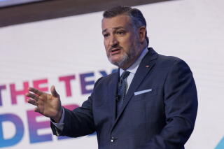 A man with a graying beard, in dark suit and tie, gestures with his hand while speaking