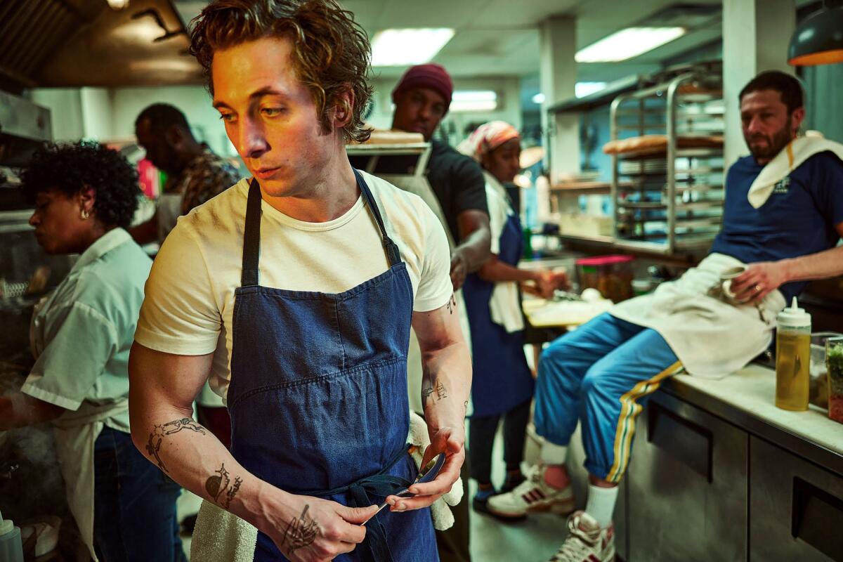 Jeremy Allen White wears a white tee and blue apron in a restaurant kitchen on the show "The Bear"