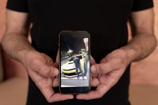 A pair of hands holding a cellphone showing a photo of a person standing in front of a white car