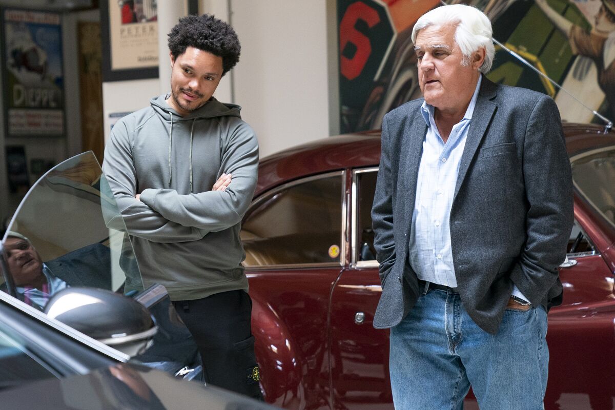 Trevor Noah and Jay Leno stand between two classic cars.