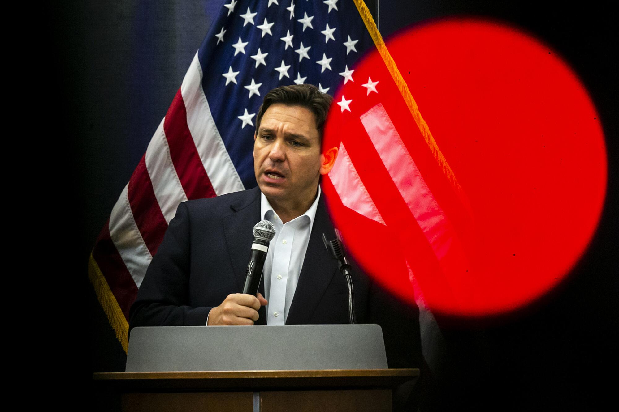 Standing in front of an American flag, Ron DeSantis speaks into a microphone. 
