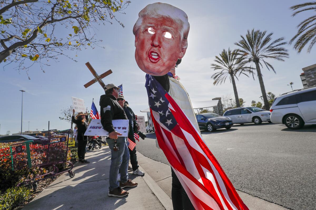 People on a sidewalk hold a cross, a flag and a Donald Trump mask.