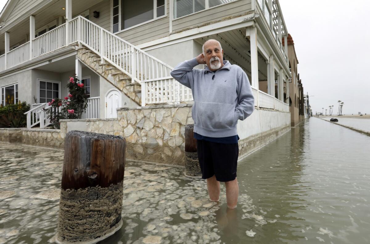 A man in basketball shorts and a hoodie stands in ocean water above his ankles next to a house near the beach