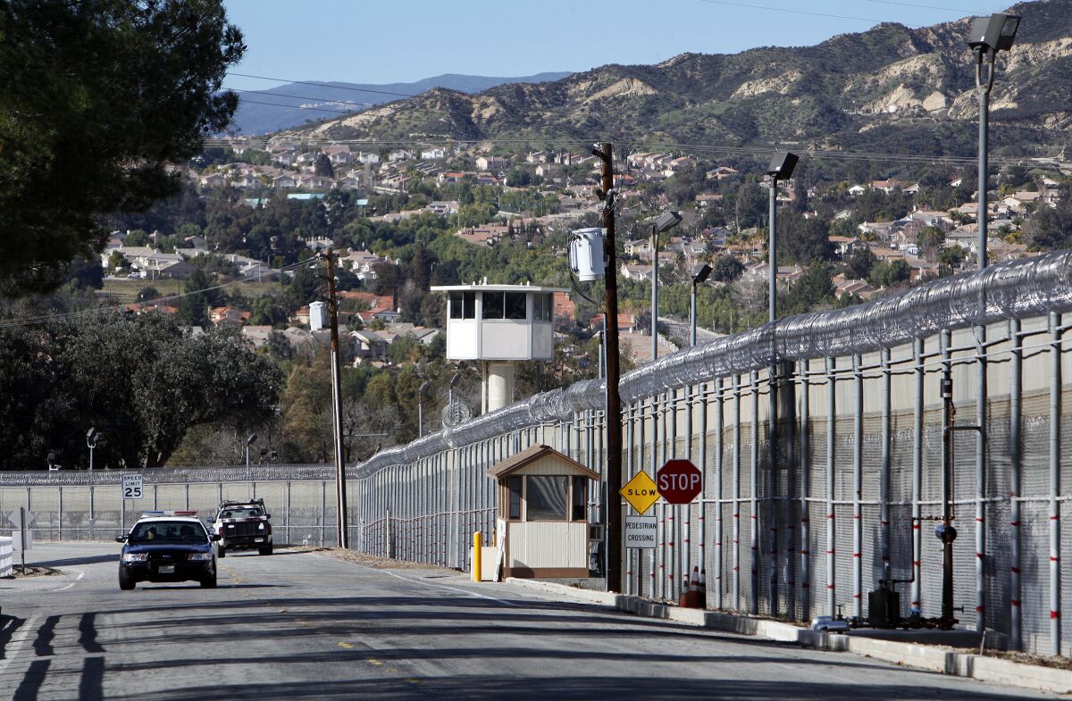 Law enforcement vehicles drive past a guard tower behind a fence topped with razor wire.