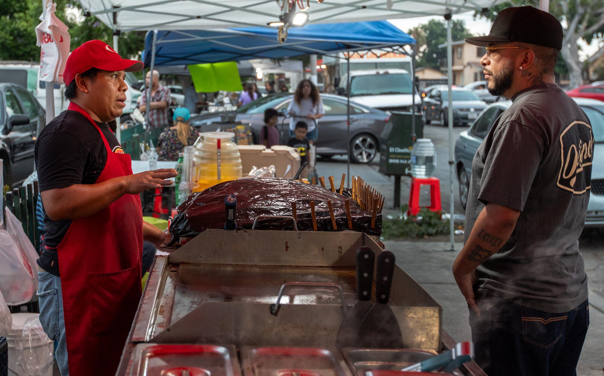 A food vendor wearing a red hat and apron speaks to a man across a griddle