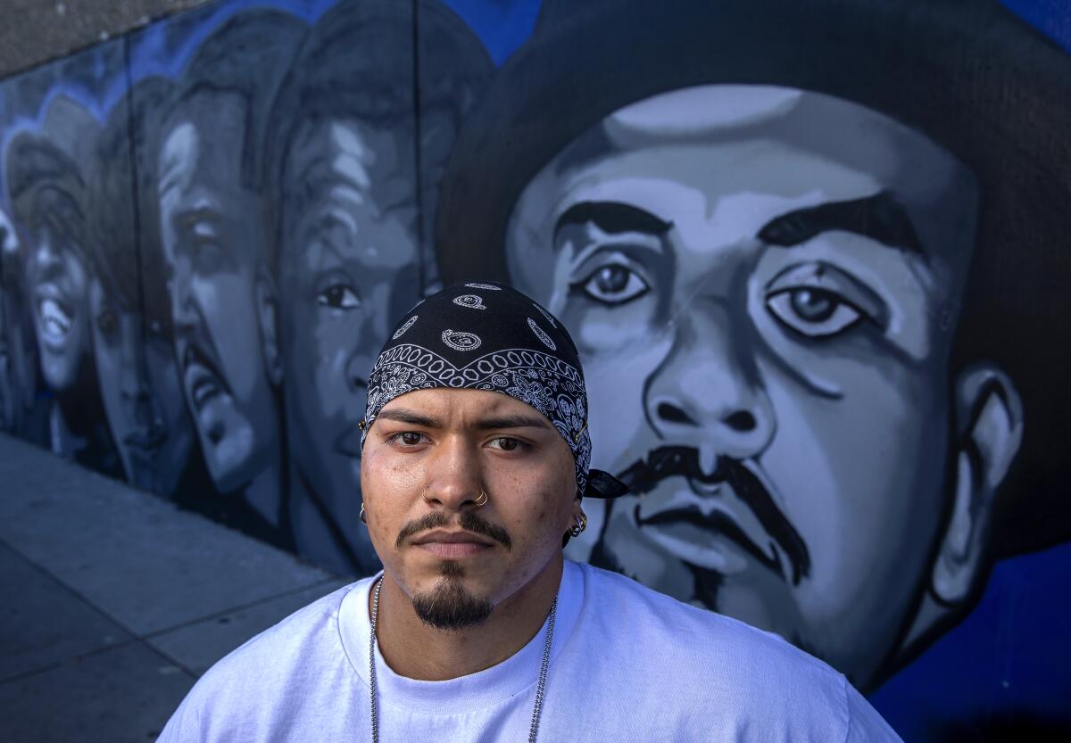 A young man with a black bandanna on his head, seen from the shoulders up in front of a mural of several large portraits