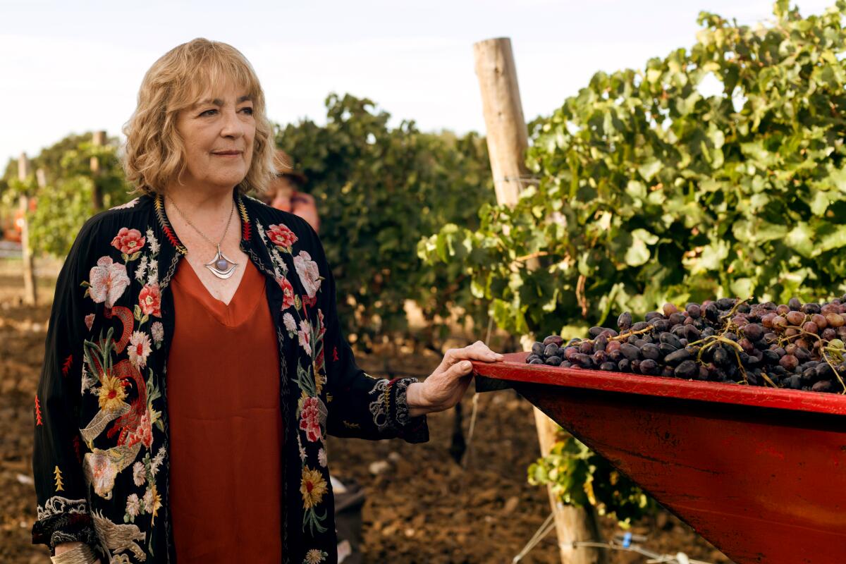 A woman in a floral jacket is standing with her hand on a cart full of grapes.