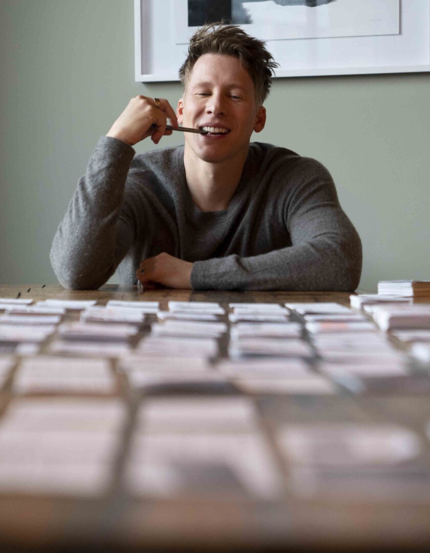 A man with a pen in his mouth sits at a table covered with index cards