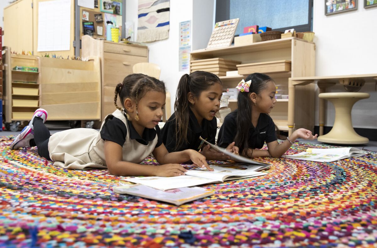 Three little girls wearing pigtails and school uniforms lie on a colorful rug reading books in a classroom