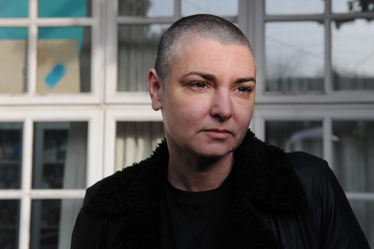 Sinéad O’Connor sitting outside in front of windows in a dark jacket.