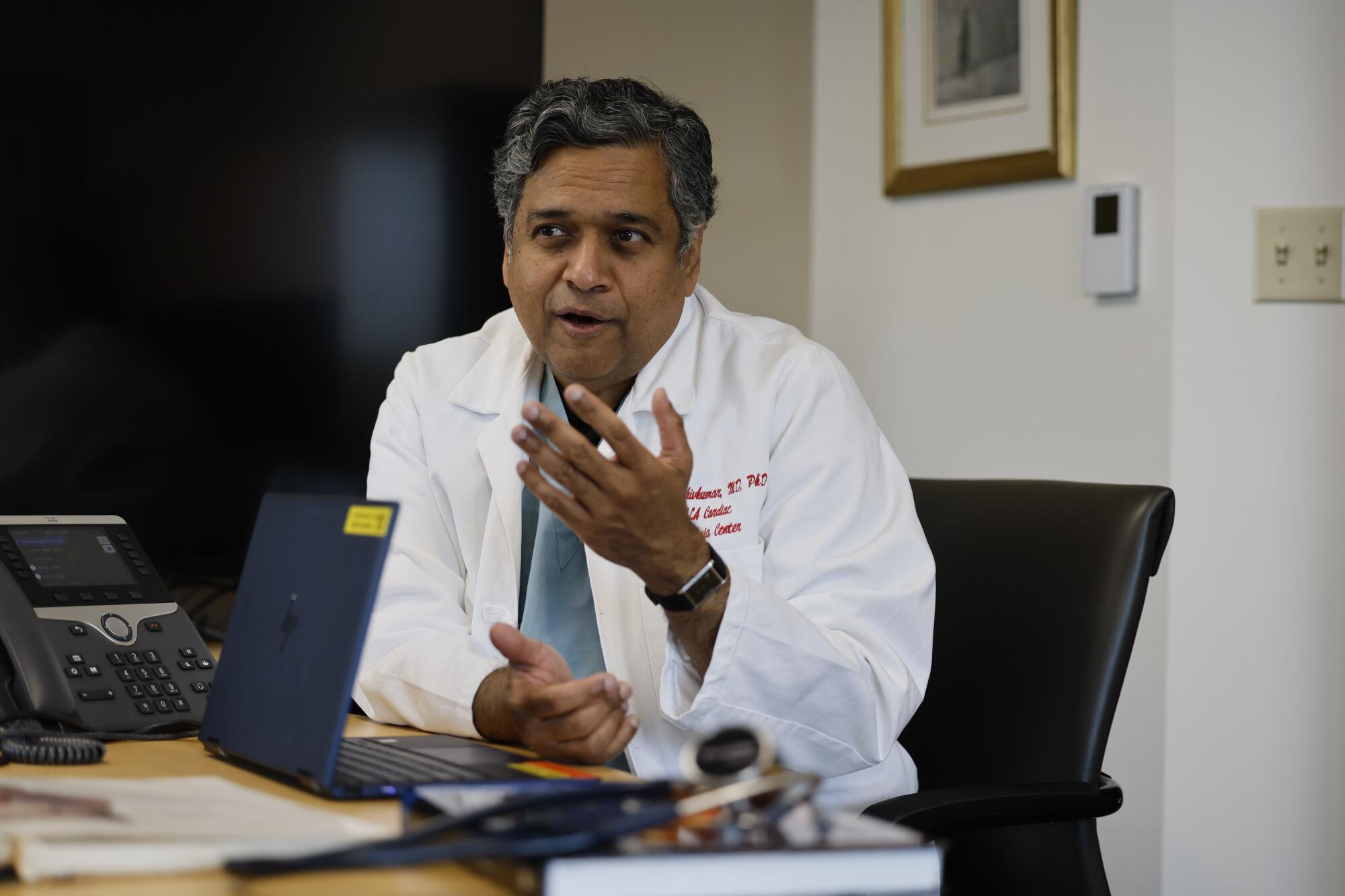 Dr. Kalyanam Shivkumar, wearing a lab coat and sitting behind an open laptop computer, gestures as he speaks.