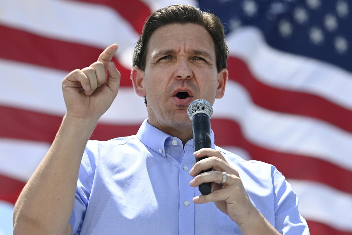 Florida Gov. Ron DeSantis speaks into a microphone and gestures in front of a huge American flag. 