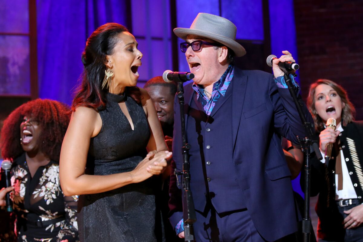 Rihannon Giddens and Elvis Costello singing into one microphone with Brandi Carlile and others singing behind them.