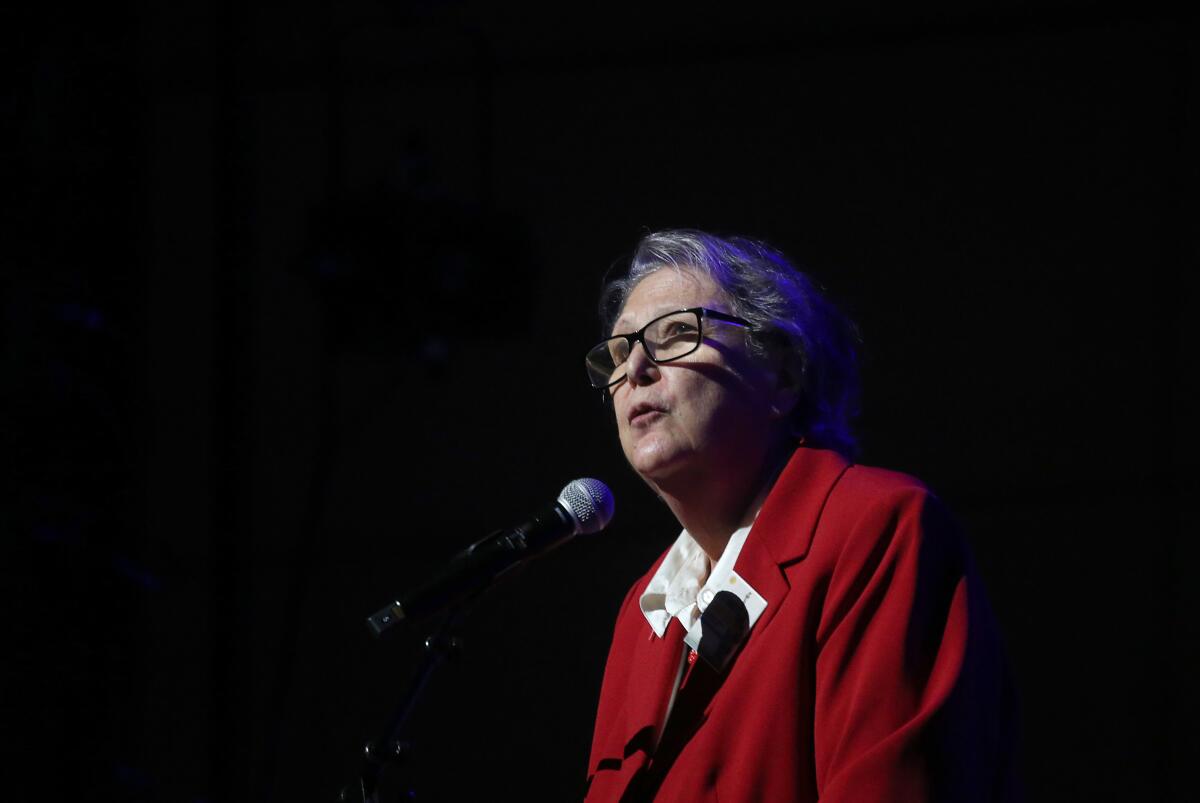 A woman in glasses and red suit jacket speaks into a microphone.
