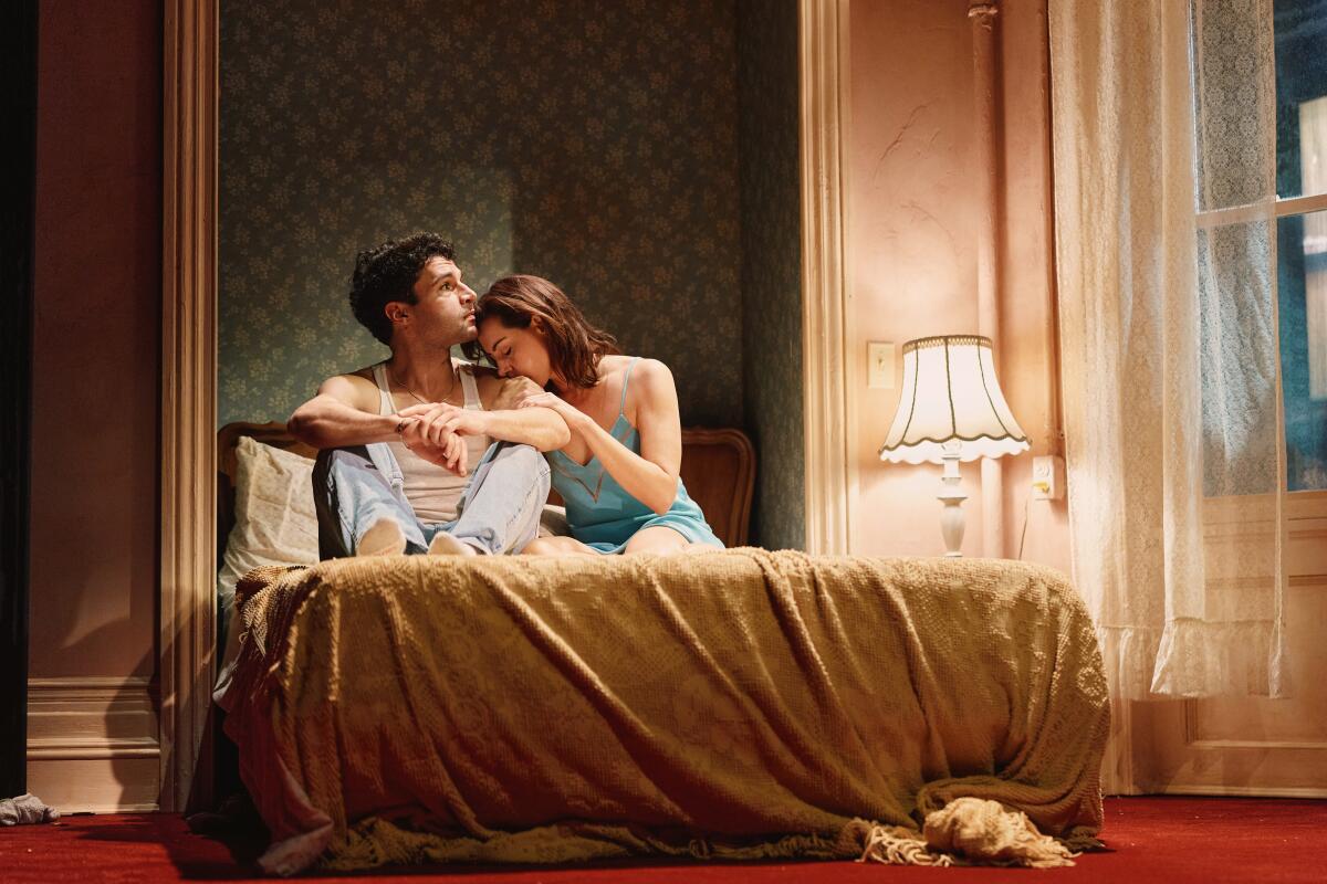 A man and a woman sit on a bed, she leaning her face against his shoulder