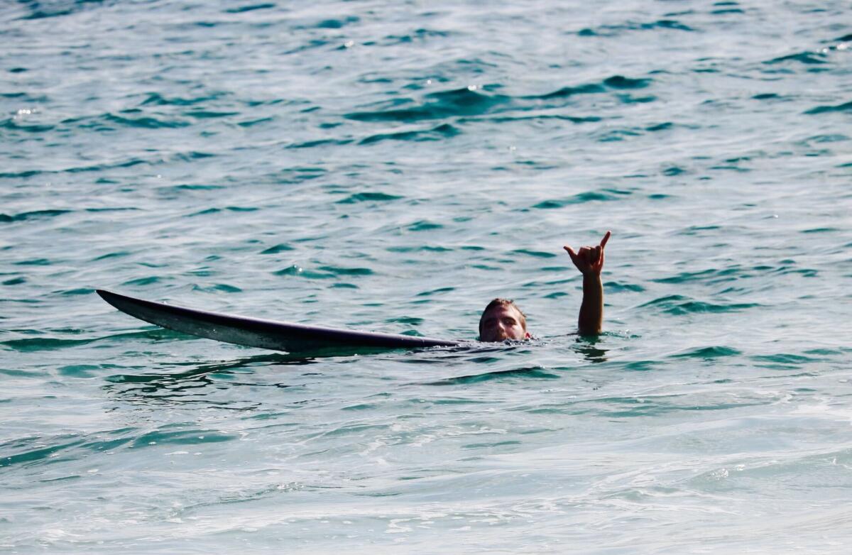 A man in deep water, holding on to a surfboard and raising one hand with his pinky and thumb extended