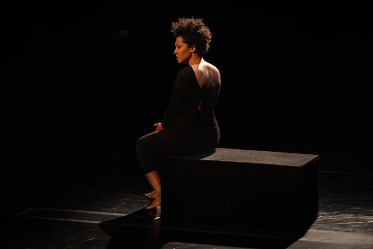 A woman onstage, seated on a black block, her body turned away from the audience