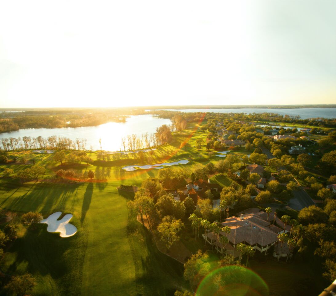 The sun reflects off a lake and shines on a golf course and trees that surround mansions.